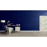 A modern room scene with a deep navy blue wall, light wood flooring, a white door, a desk, chair, lamp, and draped throw blanket.