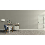 A minimalist room scene with a gray wall, light wood floor, a blue upholstered armchair with a draped throw, a wooden desk with a lamp and small accessories, a white floor lamp, and a white door on the right.