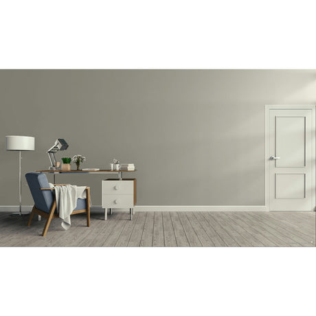A minimalist room scene with a gray wall, light wood floor, a blue upholstered armchair with a draped throw, a wooden desk with a lamp and small accessories, a white floor lamp, and a white door on the right.