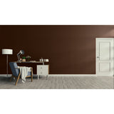 A minimalist room scene with a dark brown wall, light wood floor, a white door on the right, and a home office setup on the left featuring a blue armchair, desk, lamp, and small cabinet.