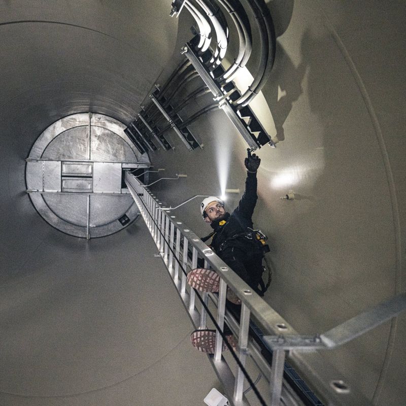 A person in a dark work outfit and white hard hat climbs a metal ladder inside a large concrete tunnel or industrial chamber, holding a flashlight and pointing upward toward overhead equipment.